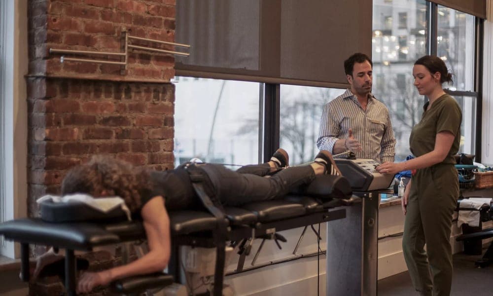 A patient lying face down on a decompression table while two clinicians review the treatment settings