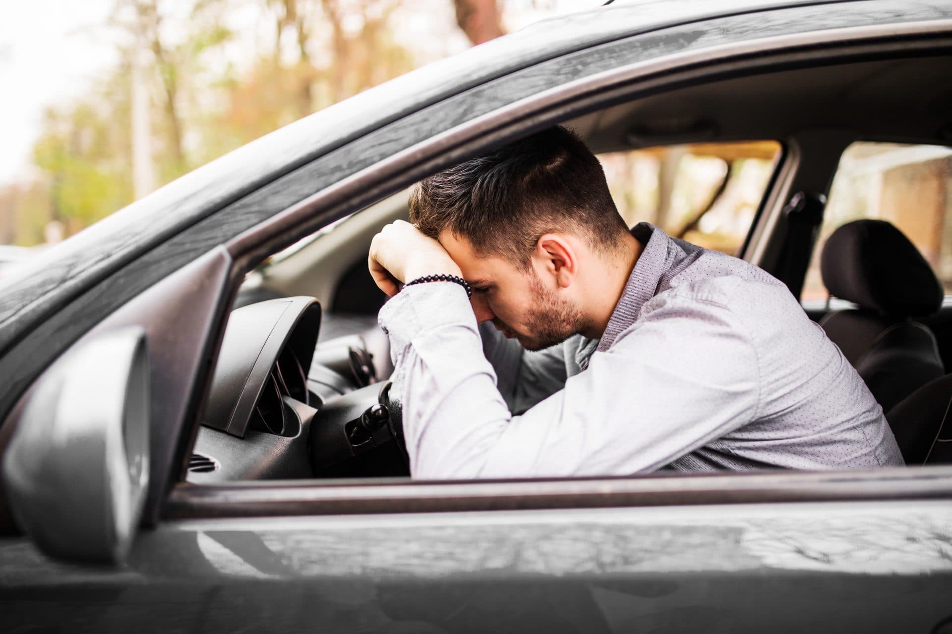 A man sitting in the driver's seat of a car resting his head against his arm on the steering wheel after a collision