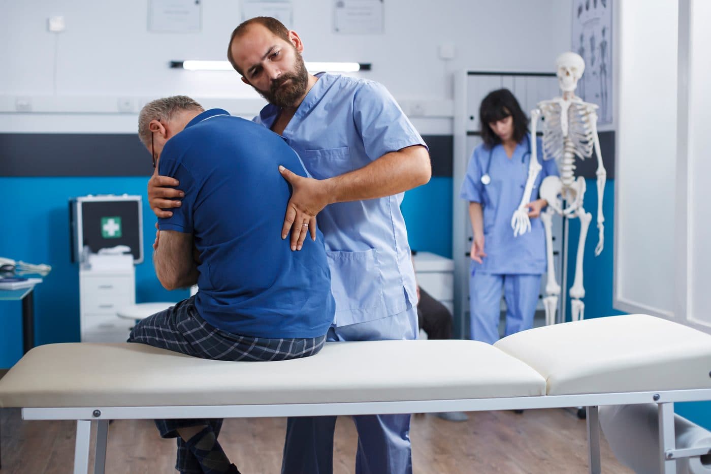 A clinician in scrubs supporting an older patient's shoulder and lower back while he sits on the edge of a treatment table
