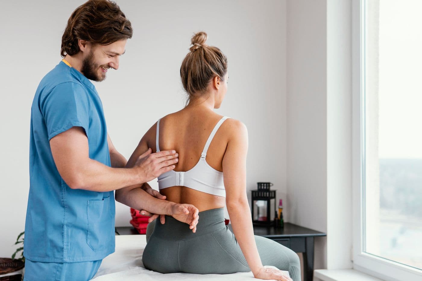 A chiropractor in blue scrubs steadying a seated patient's arm with one hand while placing the other along her mid-back