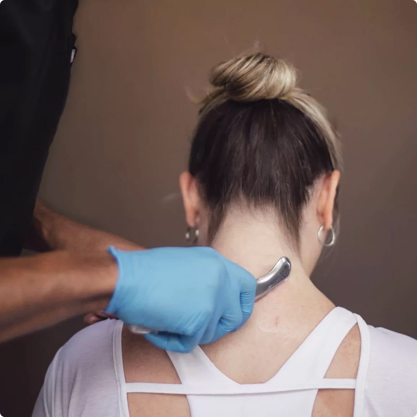 A gloved clinician drawing a stainless steel Graston instrument along the back of a seated patient's neck