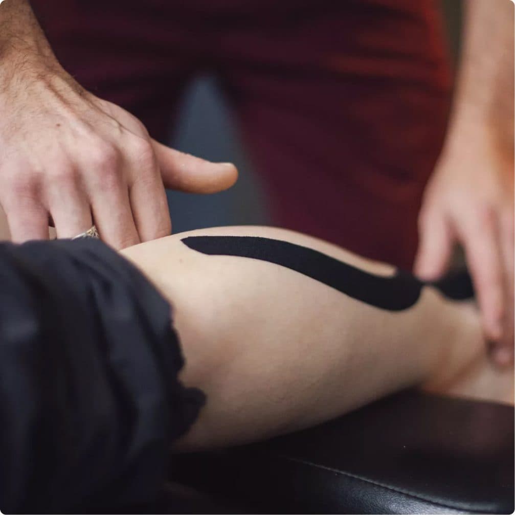 A clinician smoothing a strip of black kinesiology tape along a patient's thigh on a treatment table