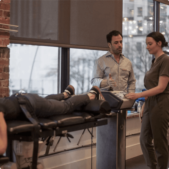 A patient lying strapped to a spinal decompression table while two Physio Logic clinicians set the treatment controls