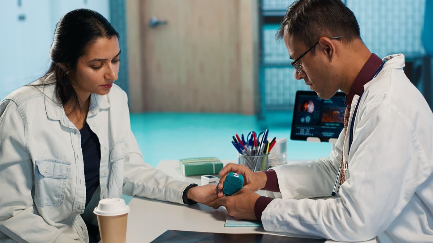 A physician in a white coat using a handheld diagnostic device on a seated patient's finger during a consultation
