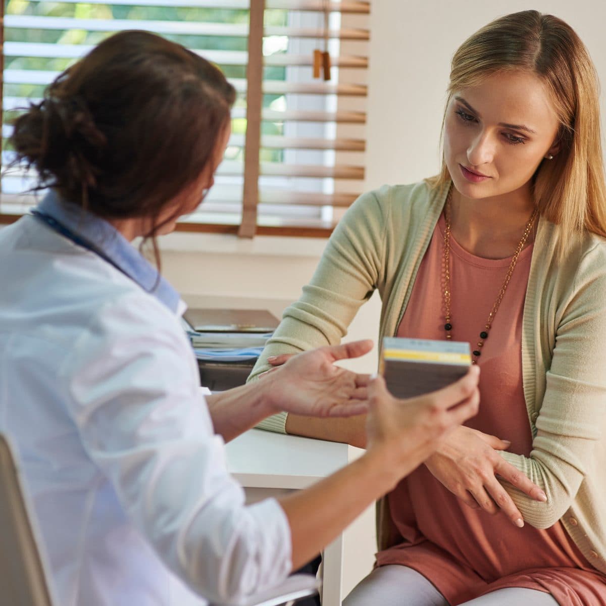 A clinician in a white coat talking a seated patient through a treatment beside a sunlit window