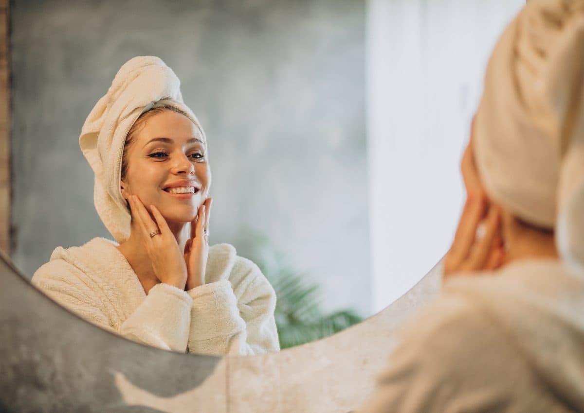 A woman in a robe with a towel wrapped around her hair applying cream to her face at a mirror
