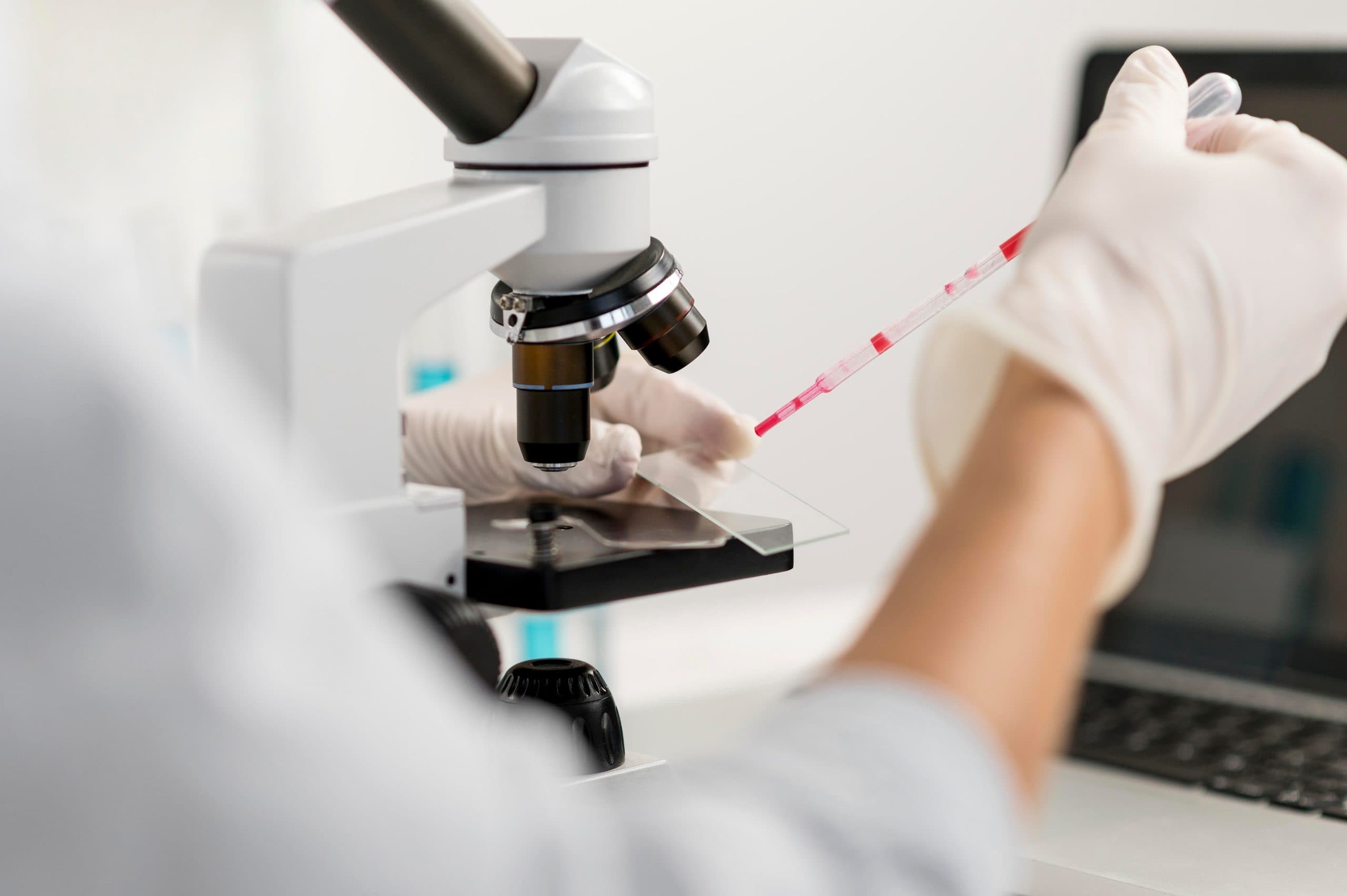A gloved hand releasing a red specimen from a pipette onto a glass slide beneath a microscope, with a laptop open on the bench behind