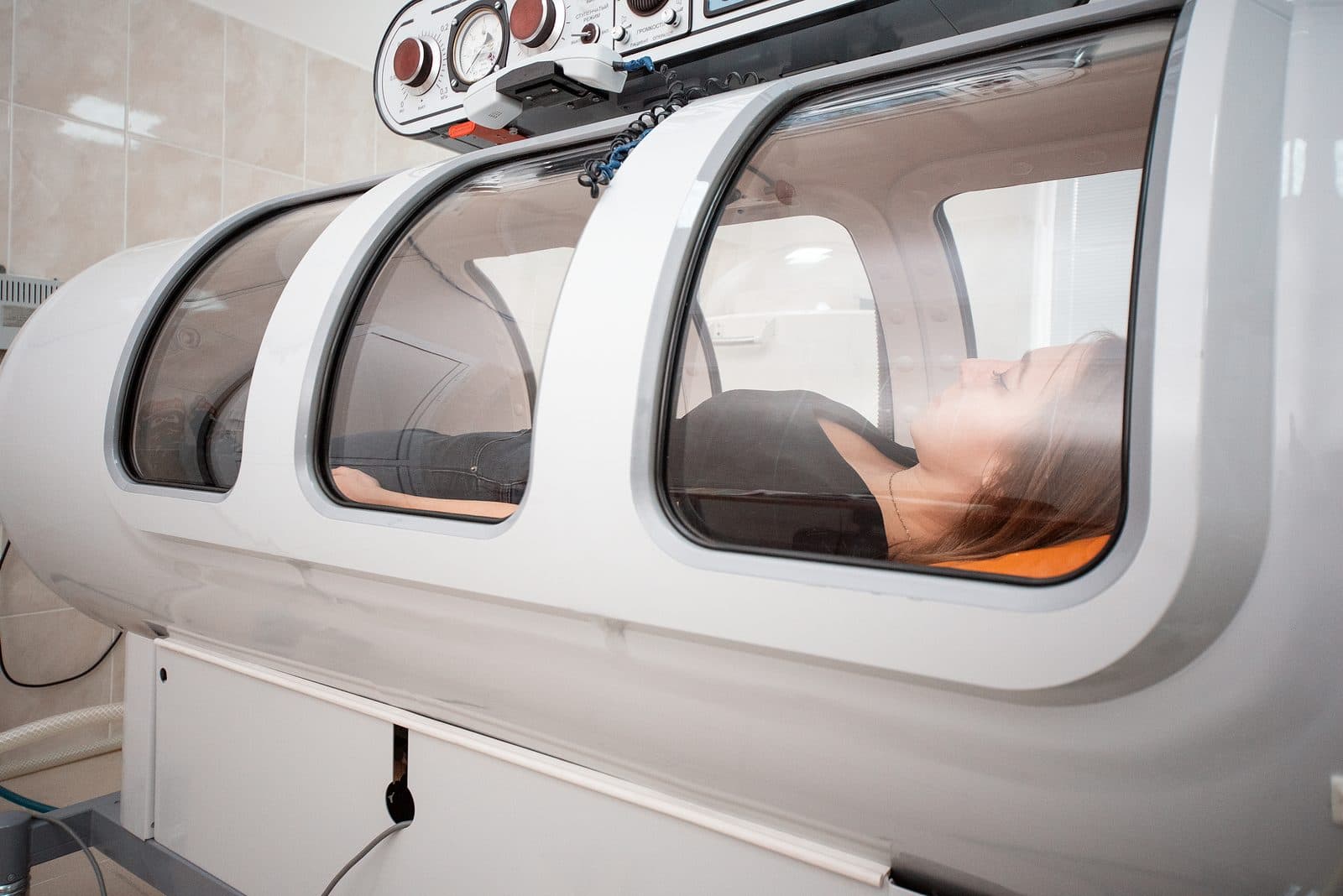 A patient resting inside a hard-shell hyperbaric oxygen chamber with three viewing windows