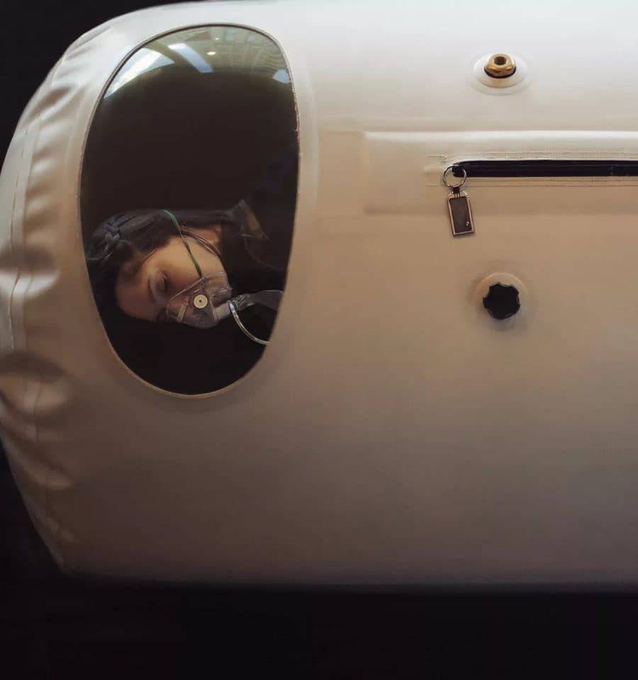 A patient wearing an oxygen mask resting inside a soft-shell hyperbaric chamber, seen through its round window