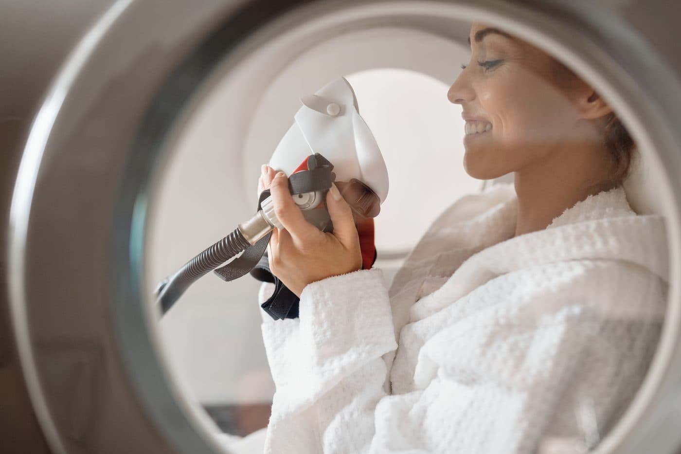 A woman in a white robe smiling as she holds an oxygen mask, framed by the round opening of a hyperbaric chamber