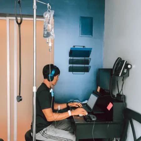 A man working at a laptop in a treatment room while an IV drip runs from a stand beside him