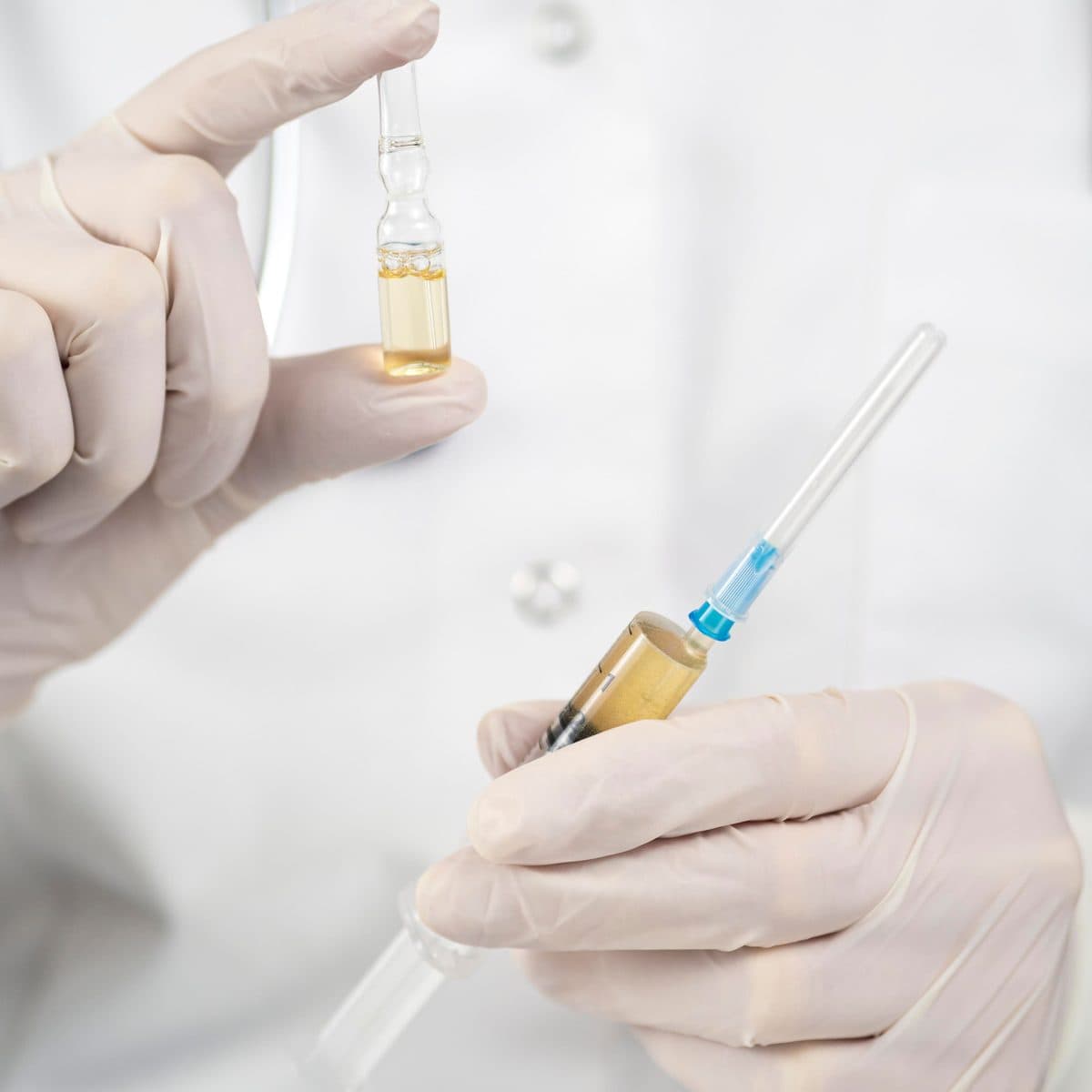 A gloved clinician in a white coat holding a glass ampoule in one hand and a filled syringe in the other
