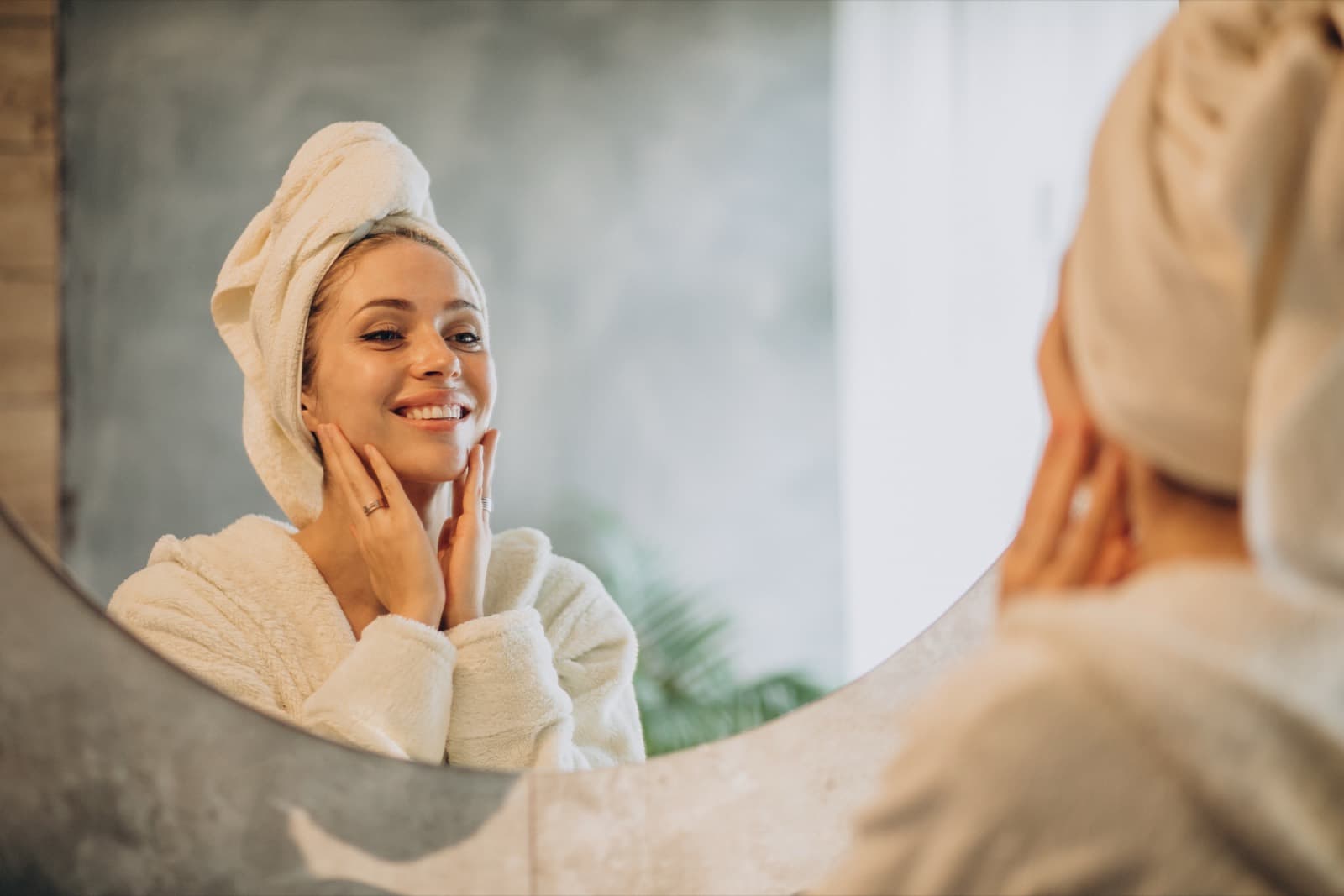 A woman in a bathrobe with a towel wrapped around her hair smiling at her reflection in a round mirror while touching her cheek