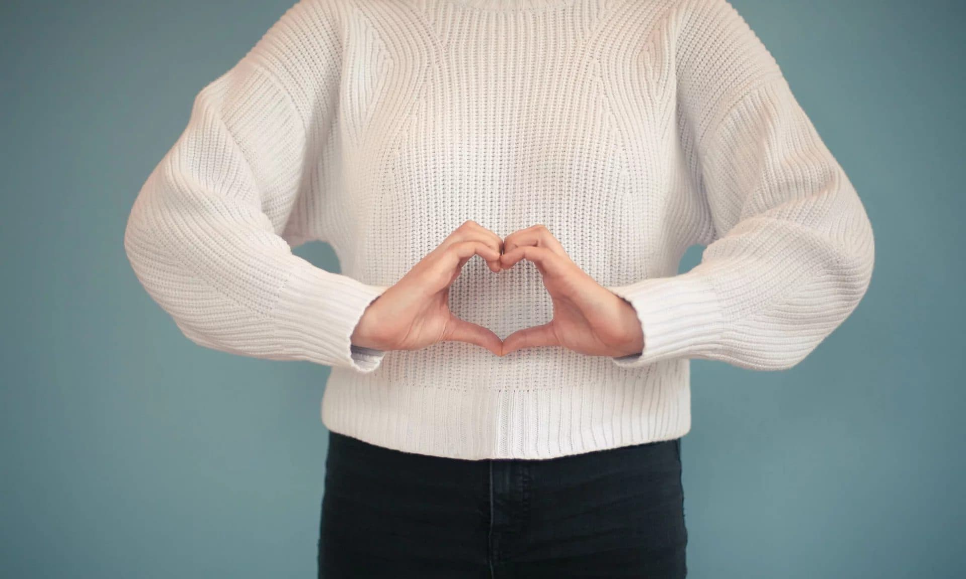A person in a cream knit sweater forming a heart shape with both hands