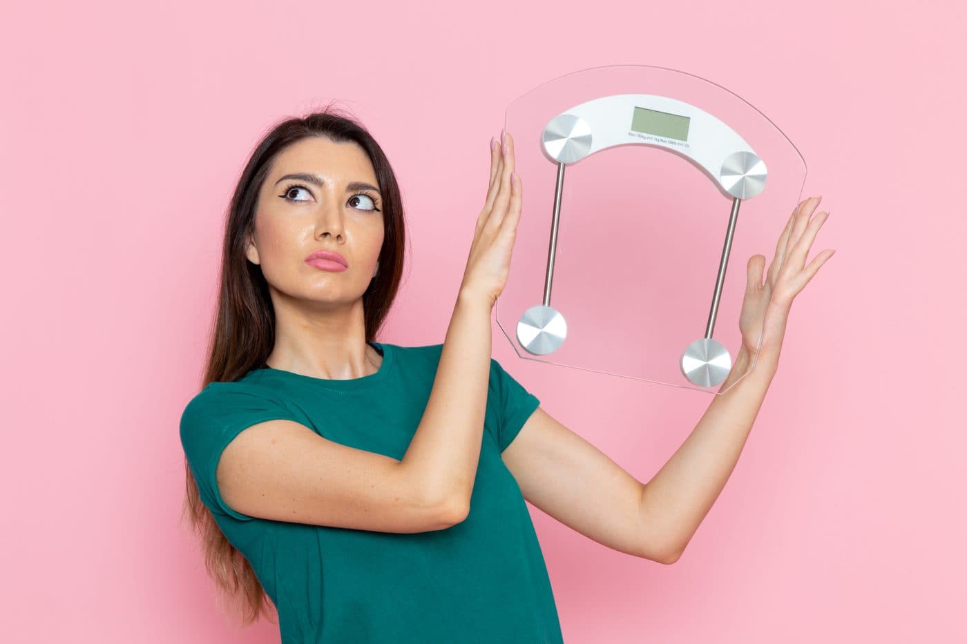 A woman raising both hands to push away a bathroom scale against a pink background