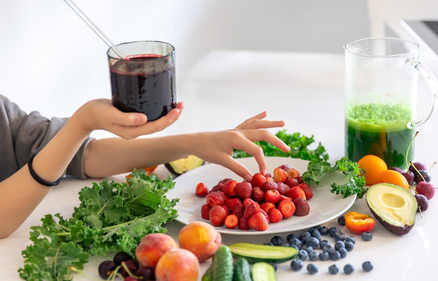 Hands holding a glass of dark berry juice above a table of fresh fruit, vegetables and a green smoothie