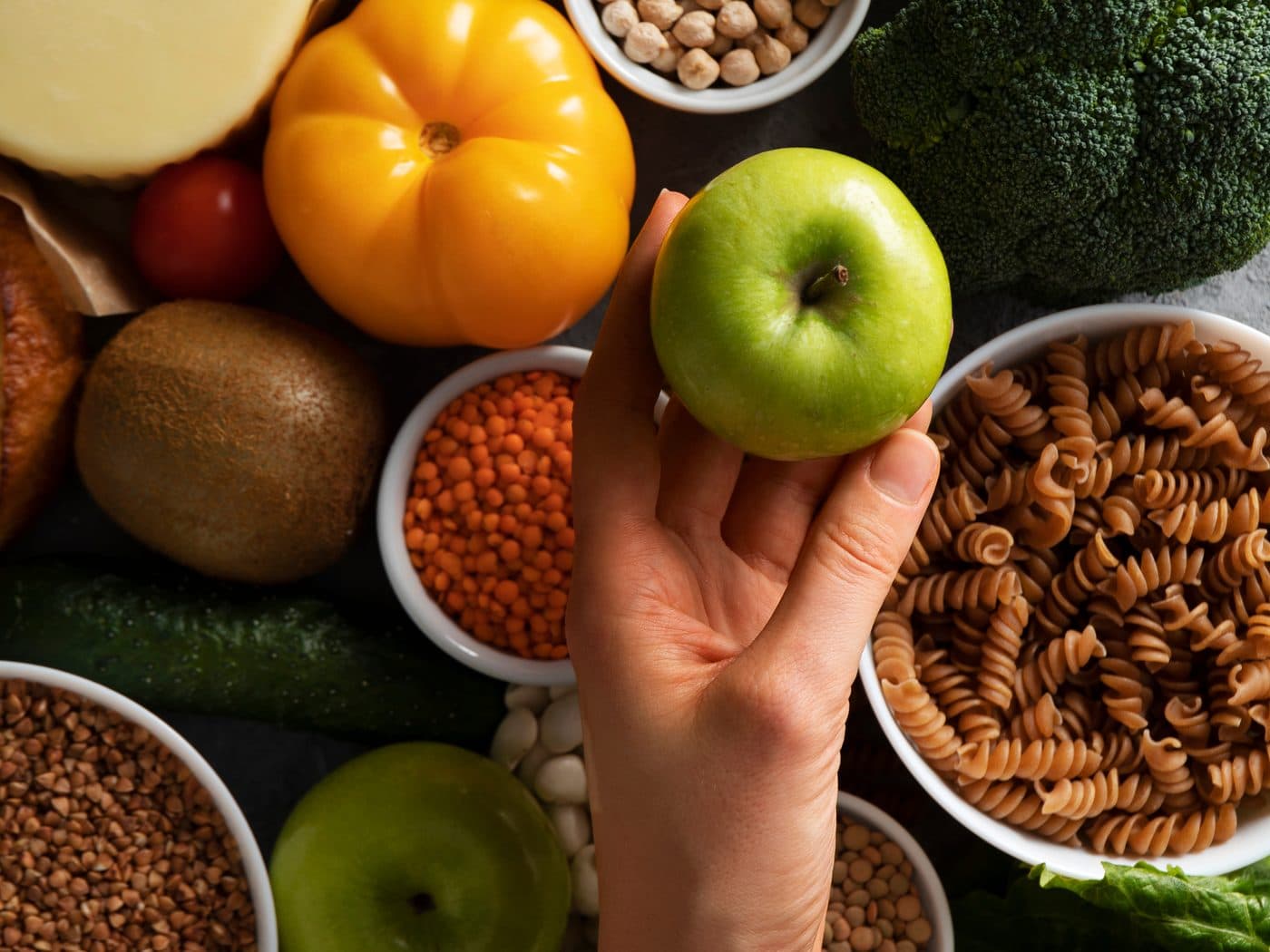 A hand holding a green apple above bowls of lentils, chickpeas, buckwheat and whole-grain pasta surrounded by fresh vegetables