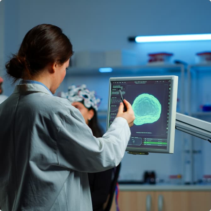 A researcher in a lab coat pointing at brain-mapping activity on a monitor while a patient wears an EEG sensor cap