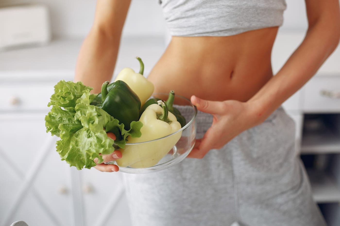 A woman in grey activewear holding a glass bowl of green peppers and lettuce in a bright kitchen