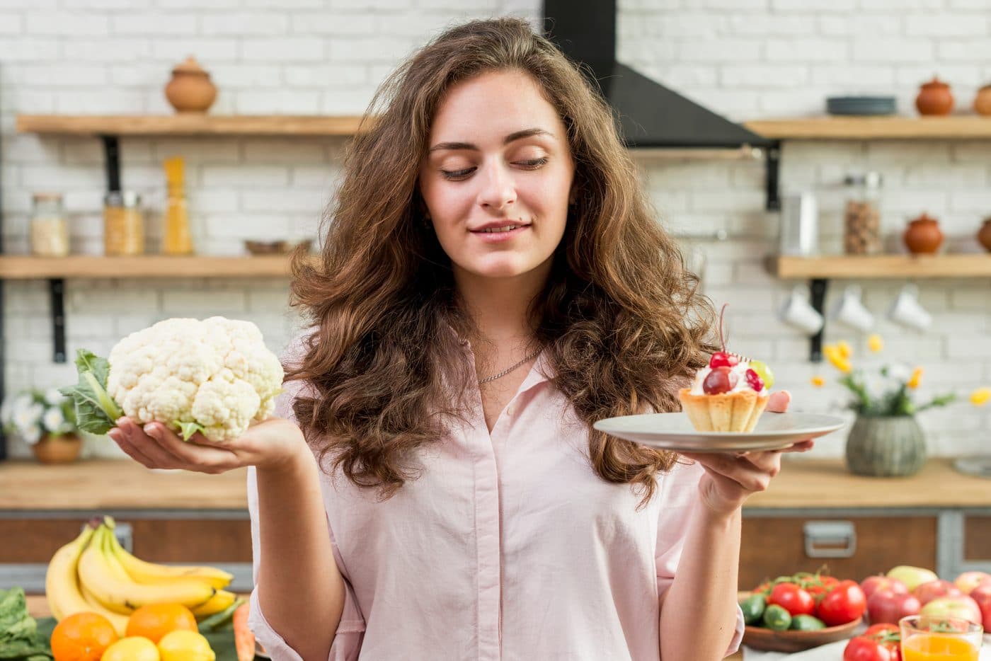 A woman in a bright kitchen weighing a head of cauliflower in one hand against a fruit tart on a plate in the other