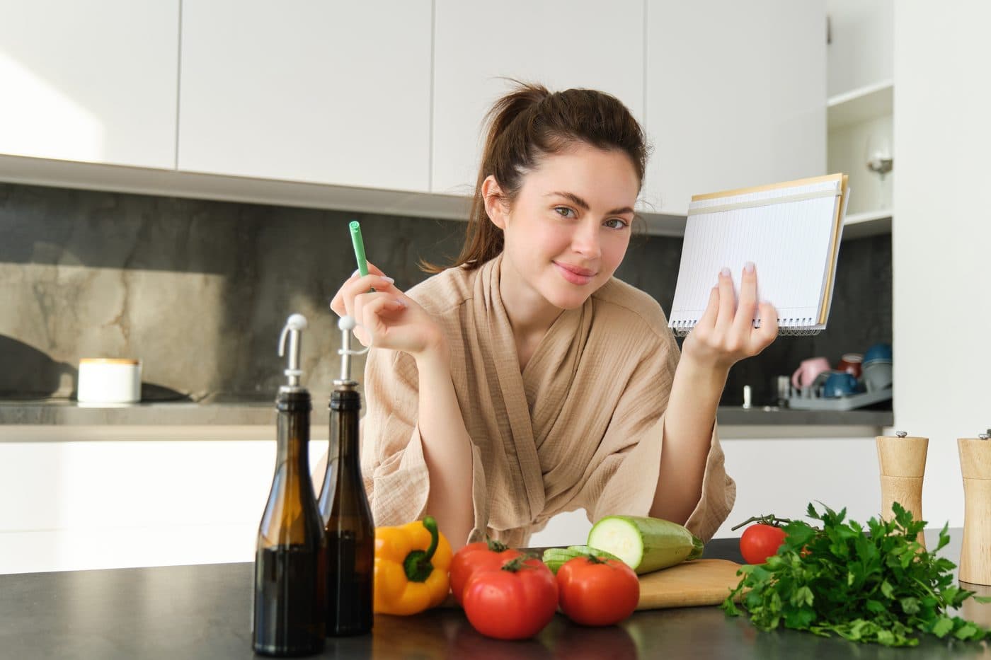 A woman leaning on a kitchen counter with a pen and open notebook beside fresh tomatoes, peppers, zucchini and herbs