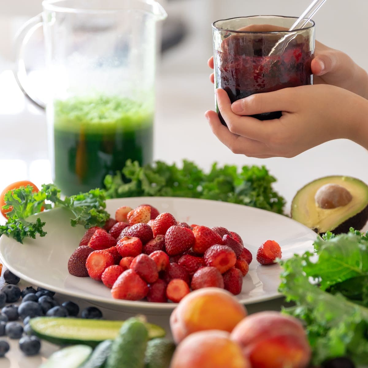 Hands lifting a glass of dark berry juice beside a pitcher of green juice, a plate of strawberries, kale, avocado, cucumbers and blueberries