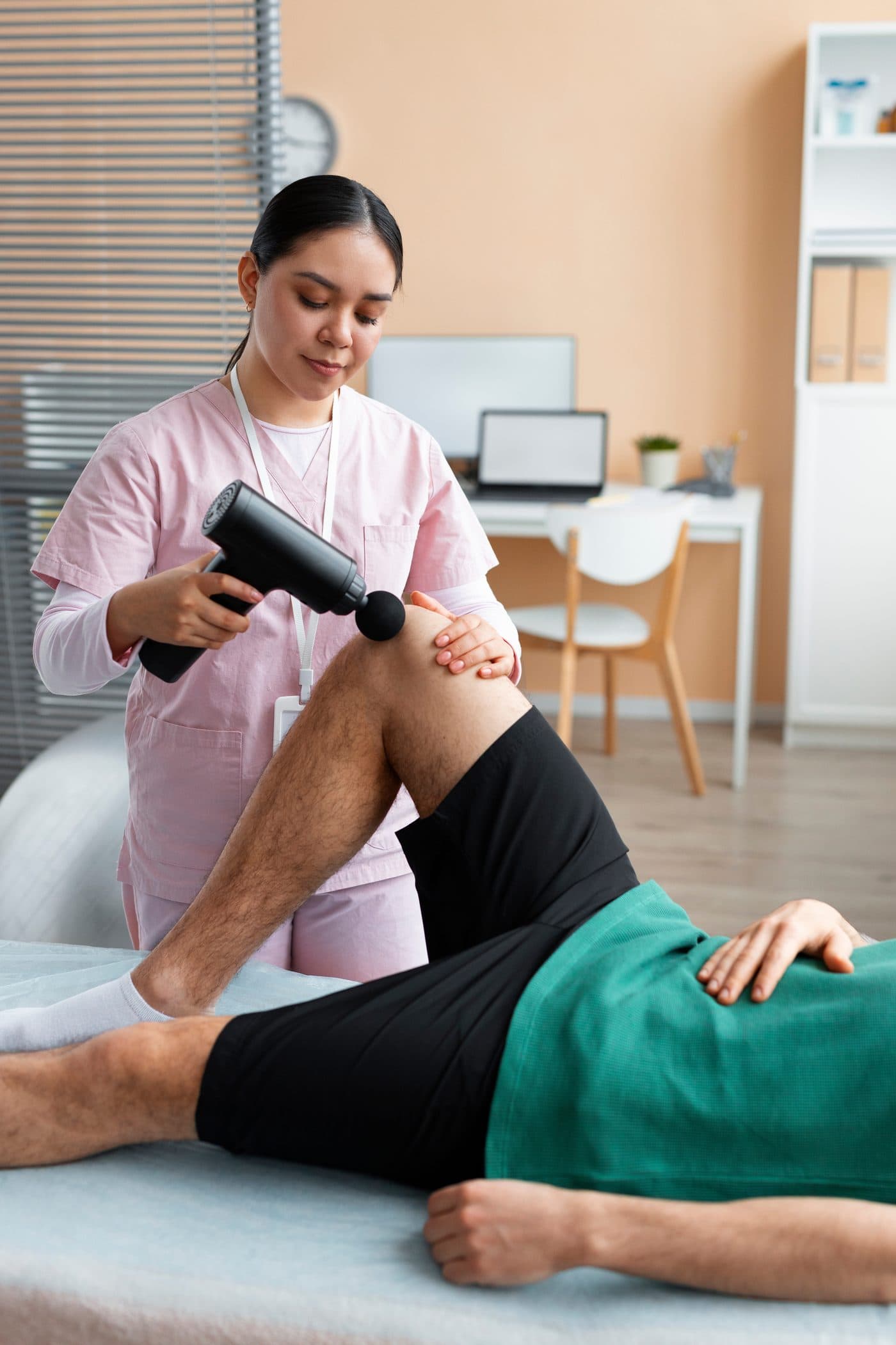A clinician using a percussive massage device on a patient's thigh while the patient lies on a treatment table