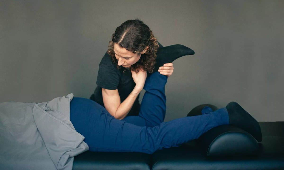 A therapist kneeling beside a side-lying patient, guiding the patient's arm across the chest during a stretch