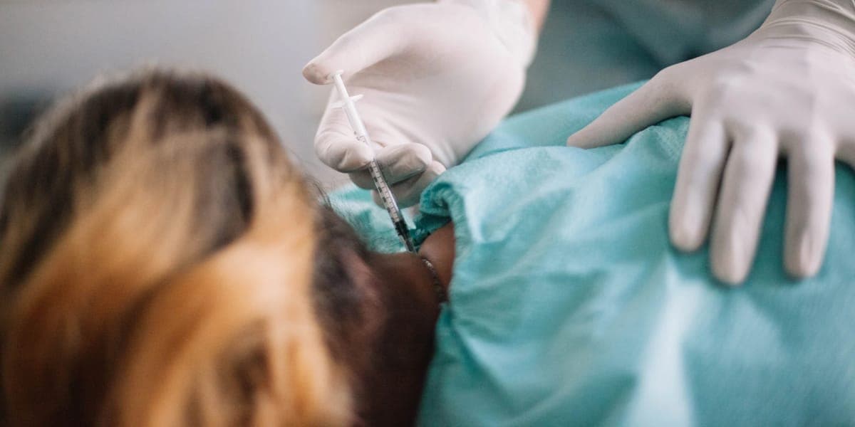 A gloved clinician administering an injection at the base of a draped patient's head