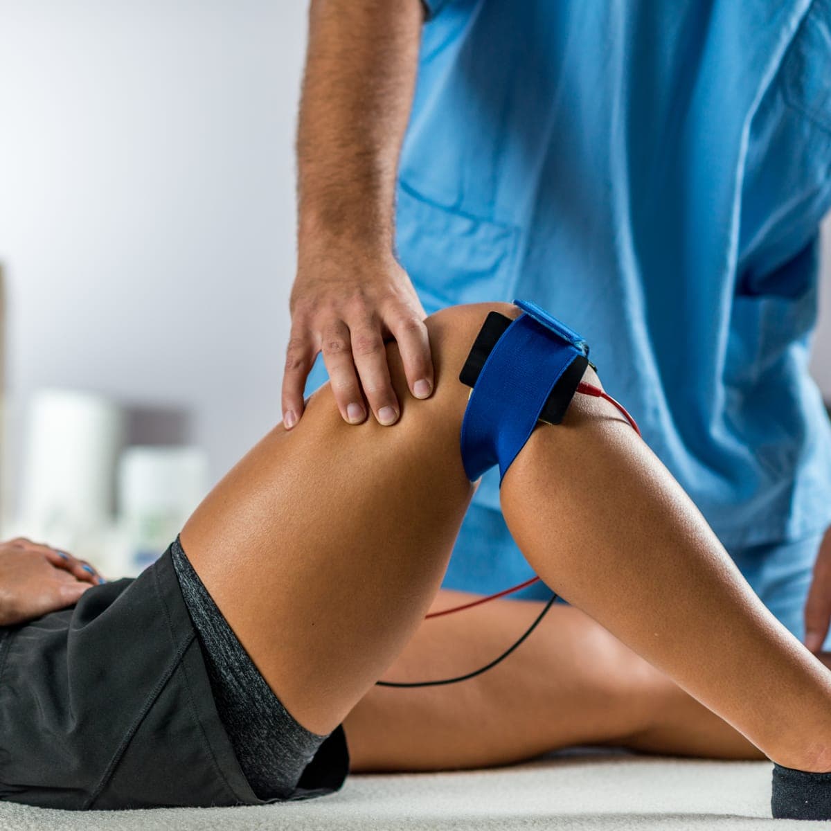 A clinician in blue scrubs steadying a patient's knee while an electrode wrap is strapped around it