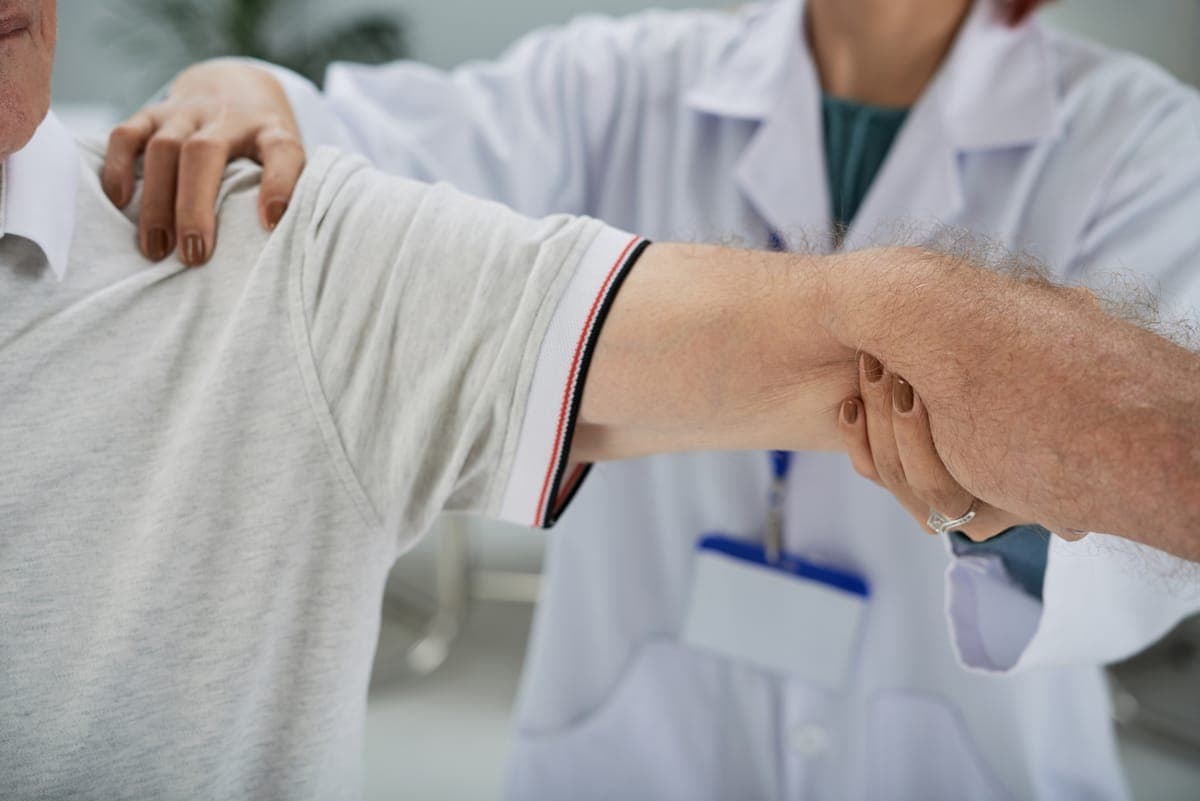 A physician in a white coat guiding an older patient's arm through a shoulder range-of-motion test