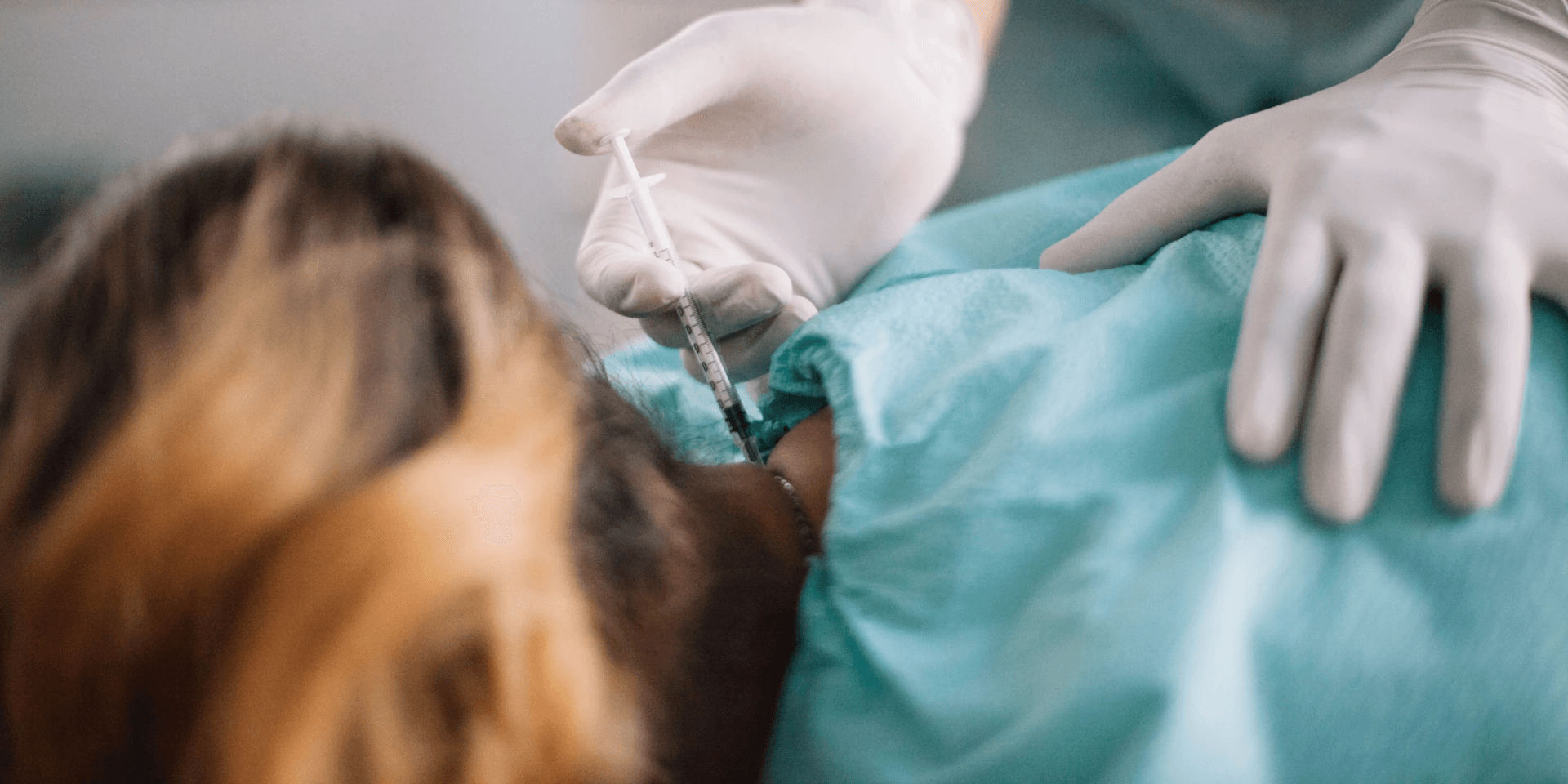 A gloved clinician steadying a draped patient and guiding a fine syringe into the base of the neck
