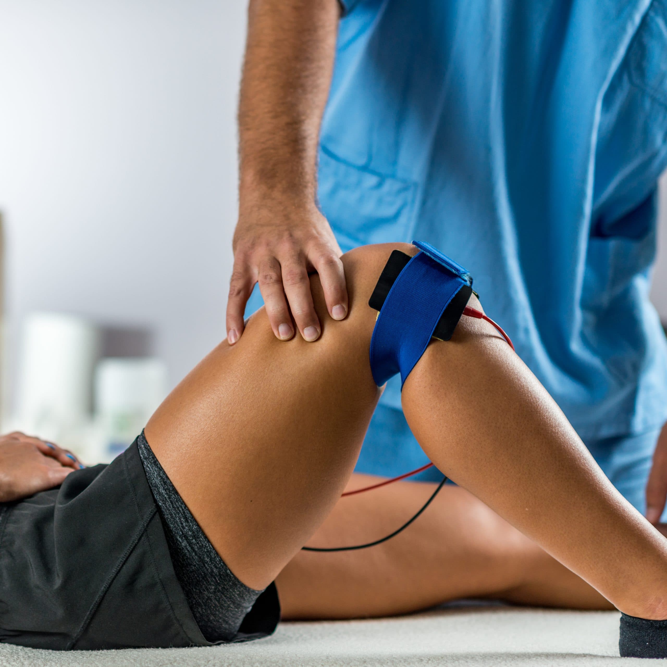 A clinician in blue scrubs steadying a patient's knee fitted with an electrotherapy strap and electrodes