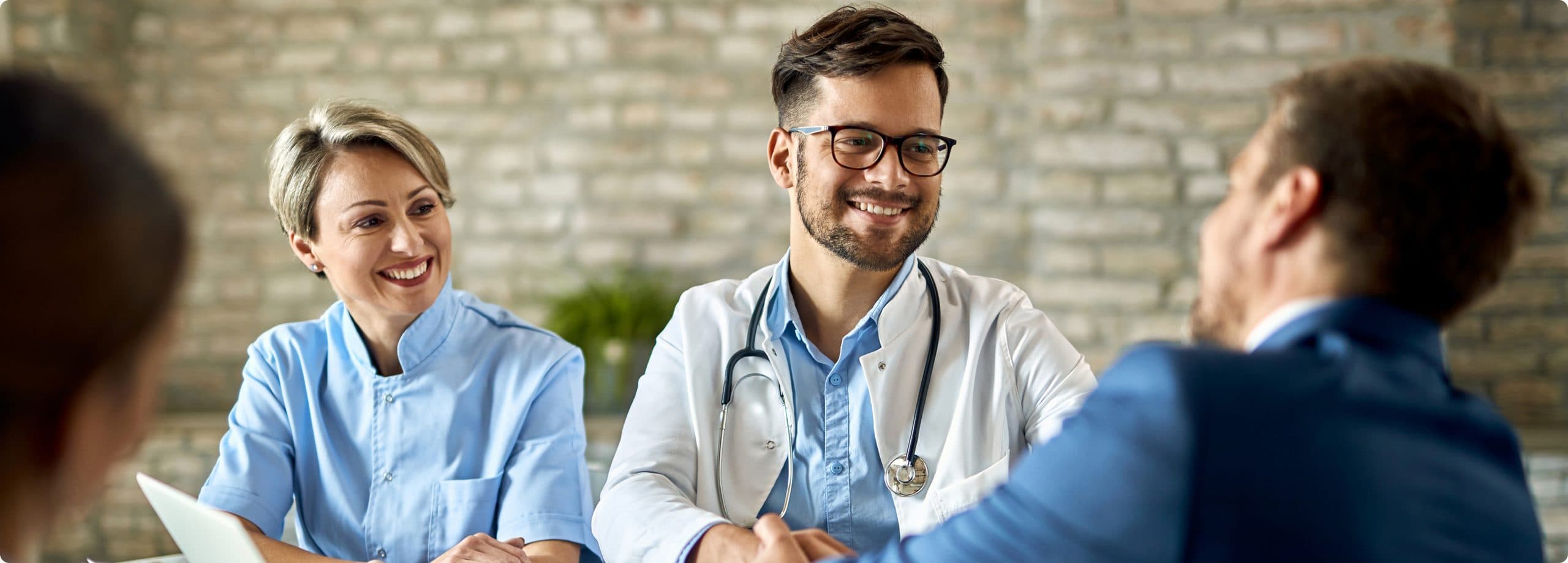 A physician and a nurse smiling as they shake hands with a patient across a consultation table