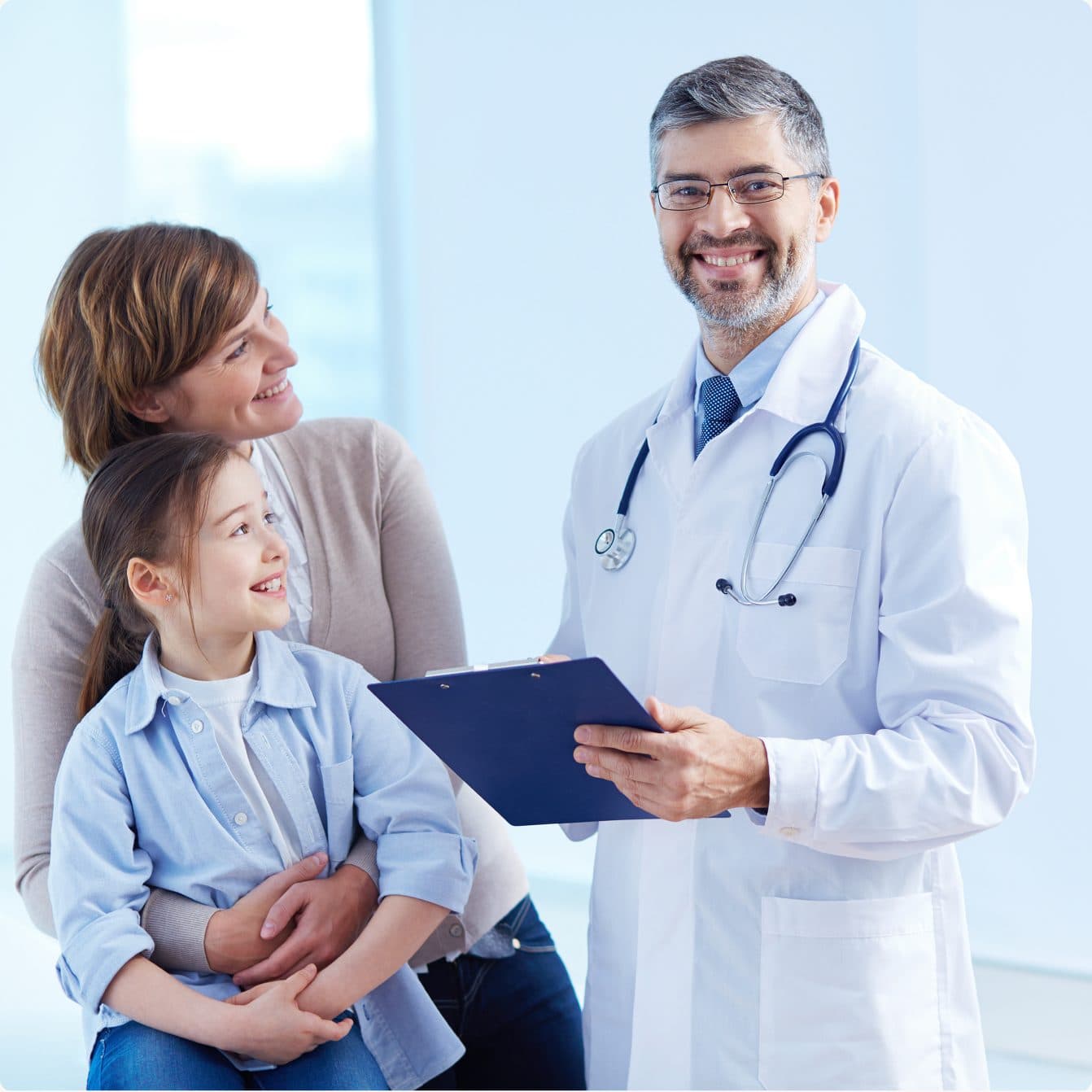 A physician holding a clipboard and smiling with a mother and her young daughter during a consultation