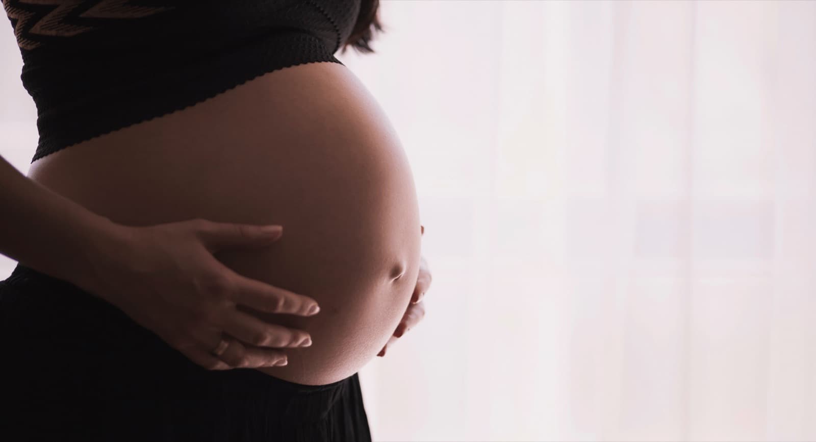 A pregnant woman resting both hands on her belly beside a sunlit window