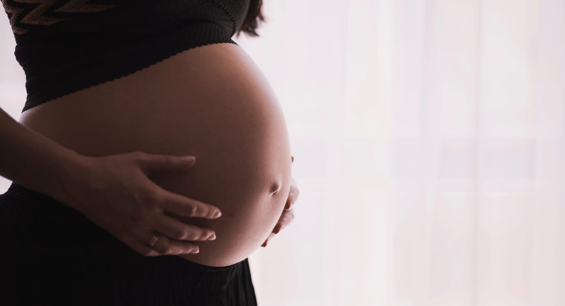 An expectant mother in a black top cradling her bare belly with both hands beside a sunlit sheer curtain