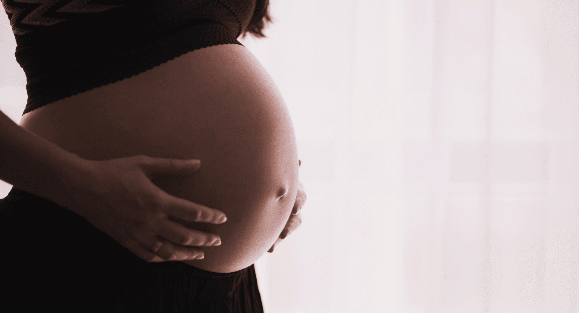 A pregnant woman resting both hands on her belly beside a bright window