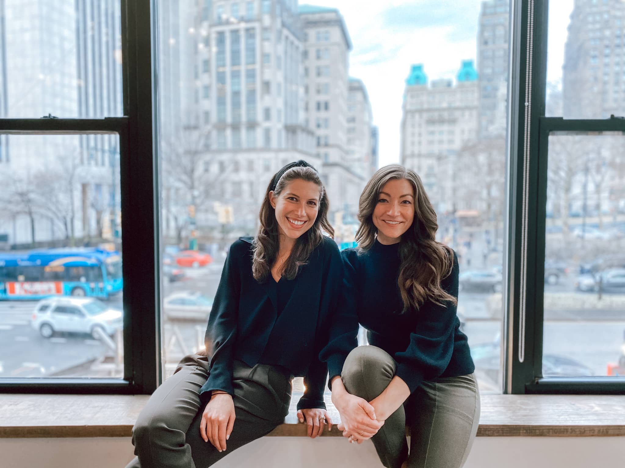The two hosts of FUNC YOU UP!, Michelle Miller and Kendra Bova, sitting side by side on a wide office windowsill, smiling, with a city street and tall buildings behind the glass