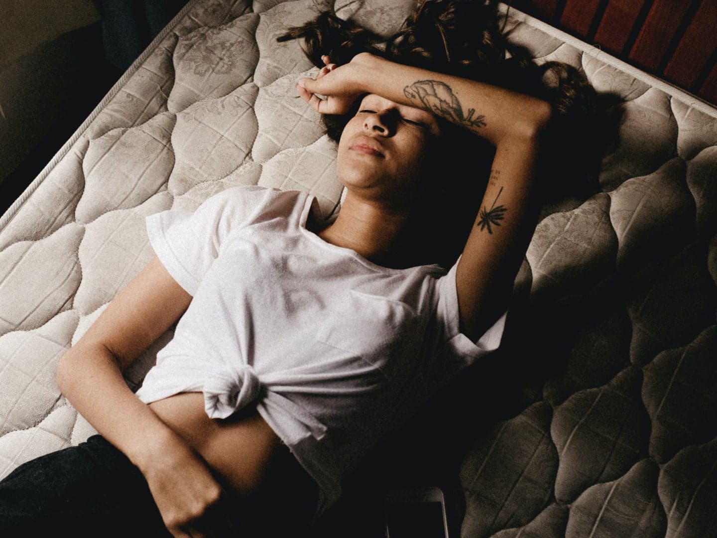 A person lying on a bed resting one arm across their eyes beside a phone