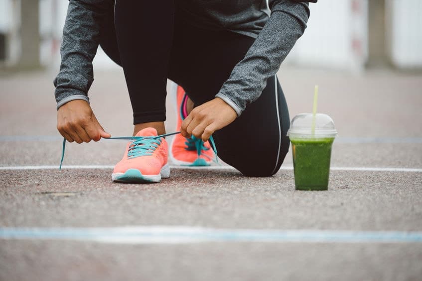A person kneeling on a running track to tie the laces of their trainers, with a green smoothie beside them